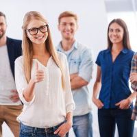 Join a successful team! Beautiful young woman showing her thumb up and smiling while group of happy young people standing on background and smiling